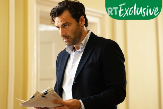 Aidan Turner as Glenn wearing a suit and standing up with a collection of papers in his hand