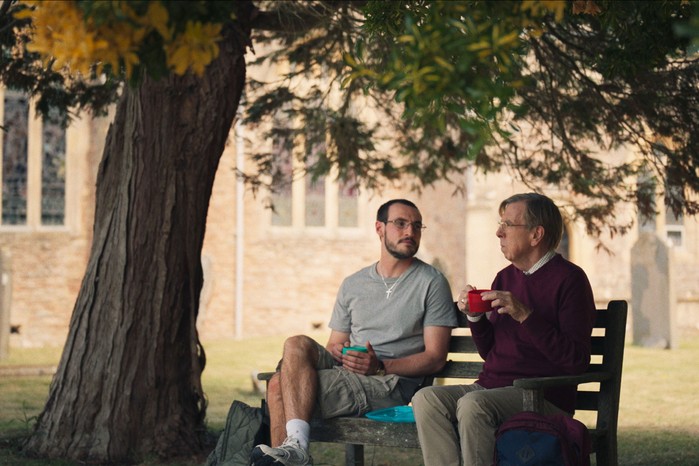 Ben and Peter sat next to one another on a bench under a tree in a churchyard drinking tea
