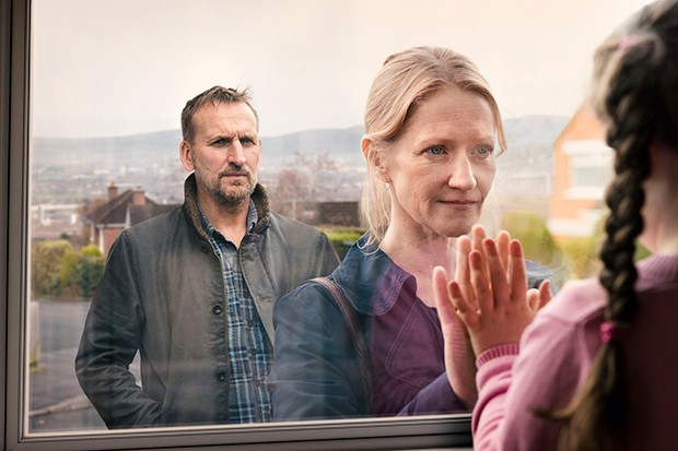 Christopher Eccleston standing behind Paula Malcomson, who has her hand pressed onto a window